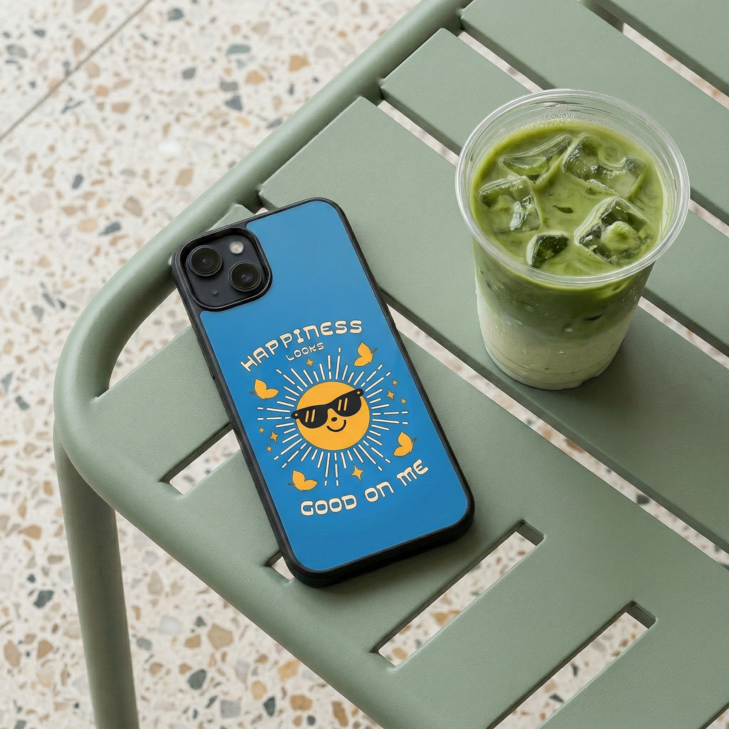 Happiness Looks Good On Me blue glass iPhone case by Spiritual Kalakaar placed on cafe table beside iced matcha drink