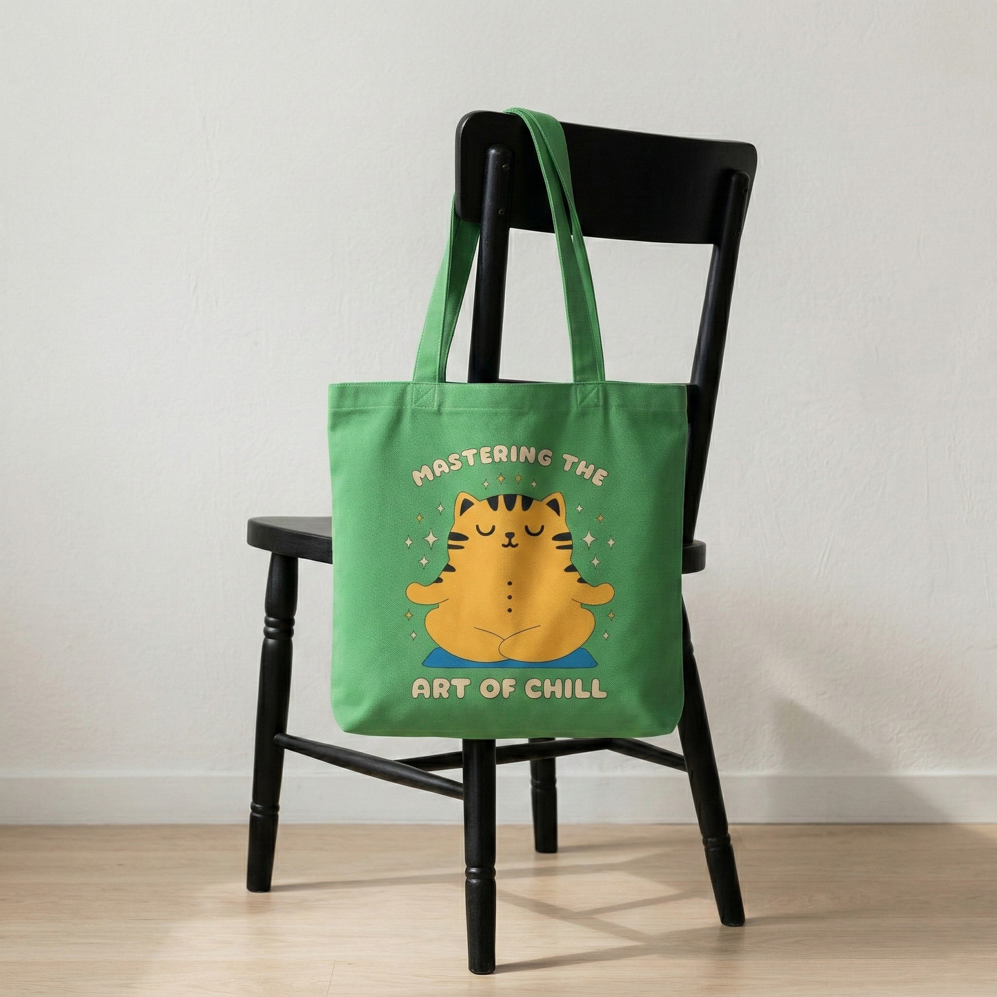 Green canvas tote bag with “Mastering the Art of Chill” meditating cat design placed on chair, cozy minimal home setting